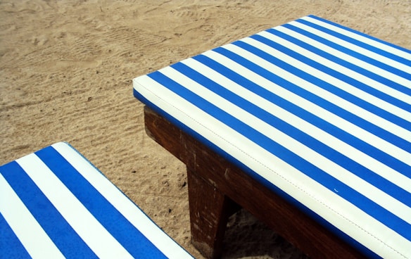 A sandy beach hosts two pieces of wooden furniture with blue and white striped cushions, evoking a nautical theme.