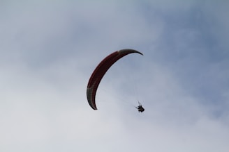 A paraglider is suspended in the sky, silhouetted against a backdrop of clouds. The parachute has a curved, red design, and the person is harnessed beneath it, enjoying a serene flight.
