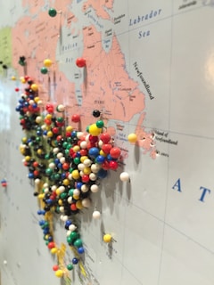 A large map is filled with colorful push pins, particularly concentrated in the region of the United States and Canada. The map details geographical areas, continental outlines, and place names, with pins marking specific locations.