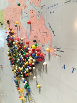 A large map is filled with colorful push pins, particularly concentrated in the region of the United States and Canada. The map details geographical areas, continental outlines, and place names, with pins marking specific locations.