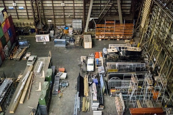 An industrial warehouse filled with various stacks and arrangements of metal beams, shelving units, and construction materials. A white truck is parked among the clutter. The scene includes colorful pallets and containers lining the walls, and the floor is mostly concrete.