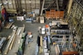 An industrial warehouse filled with various stacks and arrangements of metal beams, shelving units, and construction materials. A white truck is parked among the clutter. The scene includes colorful pallets and containers lining the walls, and the floor is mostly concrete.