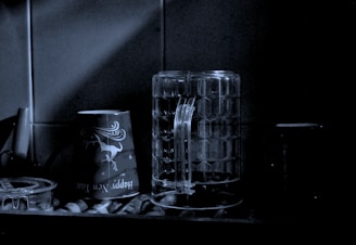 A dimly lit kitchen scene featuring a couple of cups and a large, textured glass mug positioned beside each other on a surface. The atmosphere is moody and subdued, with shadows casting over the objects. The cup on the left features festive New Year graphics. The setting appears to be quiet and still.