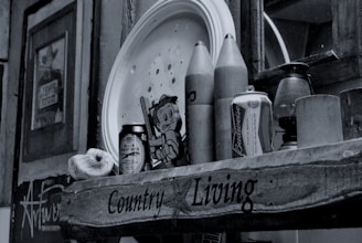 A rustic shelf displays a collection of items including a can of Budweiser, wooden rocket-shaped objects, a tin labeled 'Black Panther,' a cartoonish figure, and an antique lantern. Wall art and photos are visible in the background, contributing to an eclectic and nostalgic atmosphere.