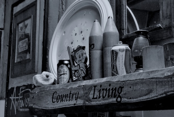 A rustic shelf displays a collection of items including a can of Budweiser, wooden rocket-shaped objects, a tin labeled 'Black Panther,' a cartoonish figure, and an antique lantern. Wall art and photos are visible in the background, contributing to an eclectic and nostalgic atmosphere.