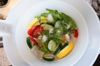 A close-up view of a white pitcher filled with slices of strawberries, cucumbers, lemons, and fresh green leaves, immersed in water with visible ice cubes floating on the surface.