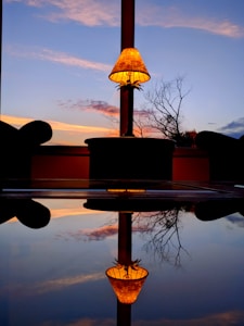A warm-lit lamp is centrally placed on a table with its reflection visible on a smooth surface, creating a symmetrical effect. The background features a large window that opens to a picturesque view of a sunset sky with soft pink and orange hues, and a silhouette of bare tree branches.