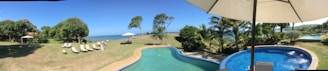 A scenic view of a coastal landscape featuring a large swimming pool in the foreground with a smaller round pool beside it. Several lounge chairs line the grassy area under the shade of tall trees. The ocean is visible in the background, framed by lush greenery and a clear blue sky.