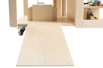 A wooden structure with a ramp leading into a compartment with shelves. The structure has caster wheels and various items such as a hammer and soda can on the shelves.