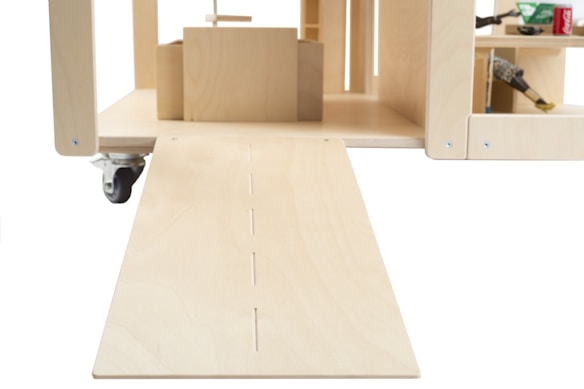 A wooden structure with a ramp leading into a compartment with shelves. The structure has caster wheels and various items such as a hammer and soda can on the shelves.