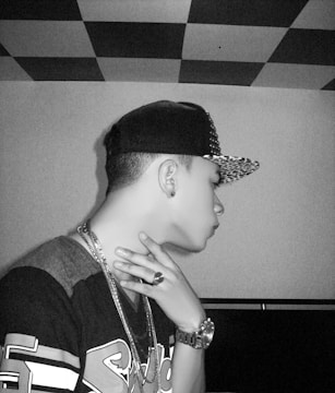 A young person is wearing a baseball cap and jewelry, including a watch and rings. The background features a checkerboard pattern on the ceiling and plain walls.