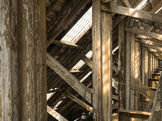 Wooden beams with peeling paint are arranged in a crisscross pattern under a sloped roof, with sunlight streaming through gaps, creating patterns of light and shadow.