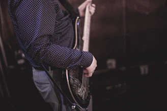 A person wearing a dark, polka-dotted shirt is playing an electric guitar. The focus is on the torso and arm, highlighting the guitar strings and picking technique.