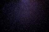 A vast expanse of a starry night sky filled with countless tiny, glittering stars scattered across a dark, inky backdrop. The density of stars increases towards the center, creating a subtle gradient from deep black to a more vibrant blueish-purple hue.