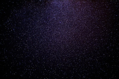 A vast expanse of a starry night sky filled with countless tiny, glittering stars scattered across a dark, inky backdrop. The density of stars increases towards the center, creating a subtle gradient from deep black to a more vibrant blueish-purple hue.