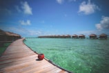 A wooden boardwalk stretches over clear turquoise water, leading to a series of overwater bungalows with thatched roofs. The sky is bright blue with scattered clouds, and a red clay pot is placed on the edge of the walkway.