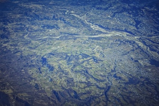 An aerial view of a vast landscape featuring a patchwork of fields, with variations in terrain, showing valleys and rivers. The land appears to be a mix of agricultural and natural areas, with roads and small buildings scattered throughout. The topography includes some elevated regions and woodland areas.