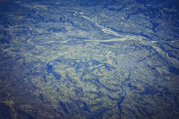 An aerial view of a vast landscape featuring a patchwork of fields, with variations in terrain, showing valleys and rivers. The land appears to be a mix of agricultural and natural areas, with roads and small buildings scattered throughout. The topography includes some elevated regions and woodland areas.