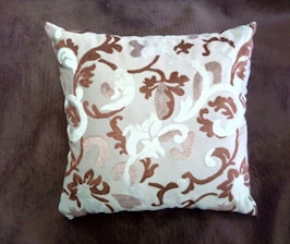 A square cushion featuring an intricate pattern of swirling, floral-like designs in shades of brown and white. The cushion is set against a dark brown background, highlighting its decorative embroidery.