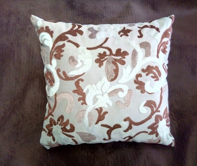 A square cushion featuring an intricate pattern of swirling, floral-like designs in shades of brown and white. The cushion is set against a dark brown background, highlighting its decorative embroidery.