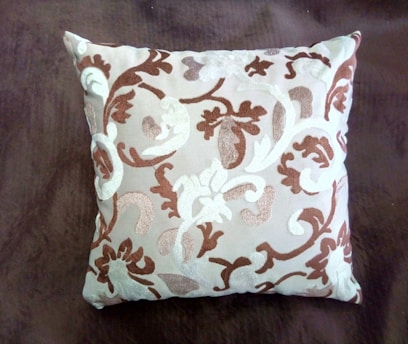 A square cushion featuring an intricate pattern of swirling, floral-like designs in shades of brown and white. The cushion is set against a dark brown background, highlighting its decorative embroidery.