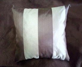 A square pillow with alternating vertical stripes of light and dark colors on a matching dark-colored surface. The fabric has a textured, slightly shiny appearance.
