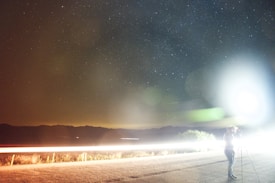 A nighttime scene depicts a starry sky above a dimly lit landscape with mountains in the distance. A bright, elongated light trails horizontally across the middle of the image, suggestive of a long-exposure photograph capturing headlights of passing vehicles. In the foreground to the right, a figure stands next to a camera mounted on a tripod, gazing upwards. A bright light source creates lens flares and illuminates part of the scene, adding a surreal quality.