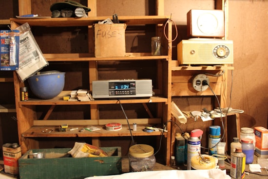 A cluttered wooden shelf in a workshop or garage containing various items, including a cardboard box labeled 'FUSE', an old radio, a digital clock showing 1:34, and various cans, jars, and small tools. There are also items like a sanding block, electrical plug extensions, and a blue basin, alongside assorted packaging materials.