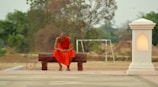 A monk dressed in an orange robe sits contemplatively on a wooden bench outdoors. Surrounding him are earthy tones, with a backdrop of green trees and sandy ground. A glowing lantern-like structure stands on his right, casting a warm light.