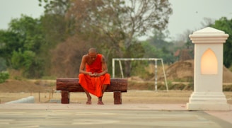 A monk dressed in an orange robe sits contemplatively on a wooden bench outdoors. Surrounding him are earthy tones, with a backdrop of green trees and sandy ground. A glowing lantern-like structure stands on his right, casting a warm light.