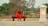 A monk dressed in an orange robe sits contemplatively on a wooden bench outdoors. Surrounding him are earthy tones, with a backdrop of green trees and sandy ground. A glowing lantern-like structure stands on his right, casting a warm light.