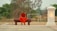 A monk dressed in an orange robe sits contemplatively on a wooden bench outdoors. Surrounding him are earthy tones, with a backdrop of green trees and sandy ground. A glowing lantern-like structure stands on his right, casting a warm light.