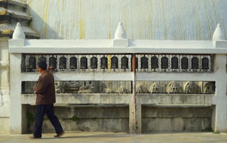 A person is walking beside a series of prayer wheels embedded in a white structure. The wheels are aligned on a metal railing, and there are carved sculptures visible below them. The architectural style suggests an environment related to spirituality or religion.