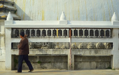 A person is walking beside a series of prayer wheels embedded in a white structure. The wheels are aligned on a metal railing, and there are carved sculptures visible below them. The architectural style suggests an environment related to spirituality or religion.