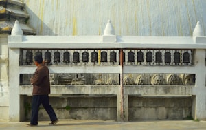 A person is walking beside a series of prayer wheels embedded in a white structure. The wheels are aligned on a metal railing, and there are carved sculptures visible below them. The architectural style suggests an environment related to spirituality or religion.