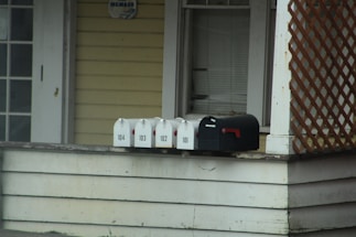 A series of mailboxes are lined up on the edge of a porch next to a house. The mailboxes are numbered 101 to 104 and are positioned on a wooden surface. The house exterior features light yellow siding and a lattice decoration. There is a dark-colored window with closed blinds above the mailboxes.