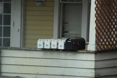 A series of mailboxes are lined up on the edge of a porch next to a house. The mailboxes are numbered 101 to 104 and are positioned on a wooden surface. The house exterior features light yellow siding and a lattice decoration. There is a dark-colored window with closed blinds above the mailboxes.