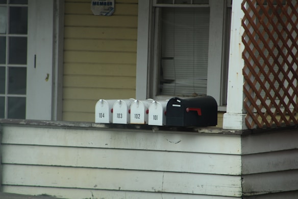 A series of mailboxes are lined up on the edge of a porch next to a house. The mailboxes are numbered 101 to 104 and are positioned on a wooden surface. The house exterior features light yellow siding and a lattice decoration. There is a dark-colored window with closed blinds above the mailboxes.