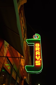 A neon sign at night displaying the word 'GRAY'S' in bright red letters outlined in green. The sign is mounted on a building with visible windows, and the scene is framed against a dark sky.