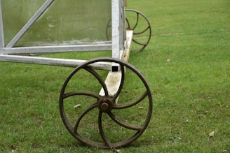 A vintage-style iron wheel attached to a metal frame rests on a lush green grassy field. The frame appears to be part of a larger structure, possibly agricultural or a garden cart.