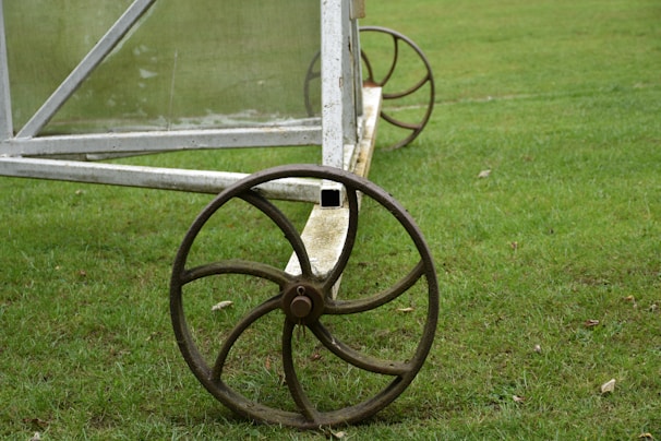 A vintage-style iron wheel attached to a metal frame rests on a lush green grassy field. The frame appears to be part of a larger structure, possibly agricultural or a garden cart.