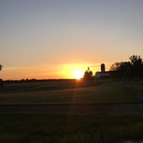 A serene landscape featuring a sunset over open fields with a silhouette of a farm building and silo visible on the horizon. The sky displays a gradient of colors from orange to blue, and trees frame the scene on the right side.