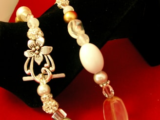 A close-up view of a decorative bracelet featuring a floral-designed clasp. The bracelet is composed of various beads including pearls, crystal-like beads, and metallic elements, all arranged on a bright red and black background.