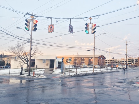 An urban intersection with traffic lights and several road signs. The pavement is wet, likely due to melting snow, and patches of snow are visible on the grass. A few bare trees are seen along with overhead utility lines. Buildings and a parking structure can be seen in the background with some people walking nearby.