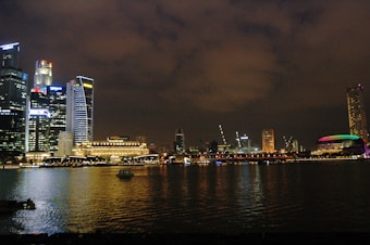 A nighttime cityscape featuring a skyline with illuminated skyscrapers reflecting on the water. The buildings have bright lights and one structure prominently displays colorful neon lights. The sky is dark with scattered clouds, adding to the overall urban atmosphere.