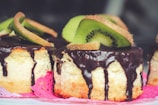 A close-up of creamy cheesecake slices topped with thick, glossy chocolate drips and garnished with fresh, vibrant kiwi slices. The cakes rest on a pink doily, creating a colorful contrast with the dessert.
