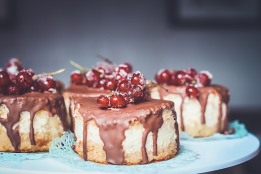 Four individual cakes topped with a generous amount of chocolate glaze and garnished with clusters of red currants. The pastries are placed on a light blue doily, creating a visually appealing dessert presentation.