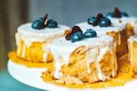 Small round cakes topped with a rich white icing, garnished with fresh blueberries and chocolate pieces, placed on a decorative yellow doily. The texture of the cake appears moist and fluffy, with the icing gently cascading down the sides.