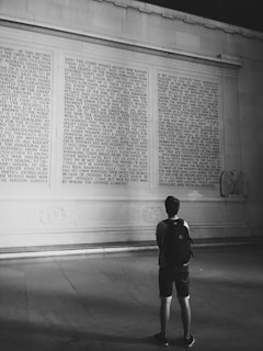 A person with a backpack stands in front of a large stone wall inscribed with a lengthy historical text. The setting appears to be indoors with dim lighting, enhancing the focus on the etched writing.