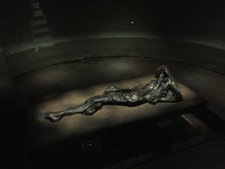 A darkly lit room features a metallic sculpture of a reclining male figure, positioned on a rectangular platform. The details of the figure's muscular form are highlighted by the dim light, casting subtle shadows. The surrounding space appears minimalistic, with a focus on the artwork.